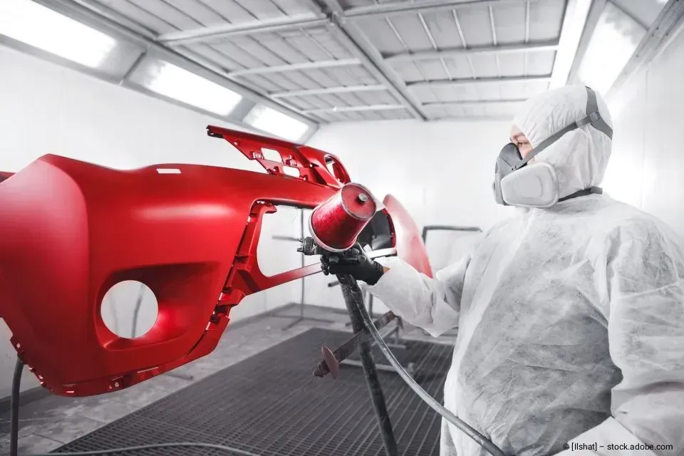 Eine Person in einem weißen Anzug besprüht in einer hellen Autolackierkabine eine rote Autokarosserie.