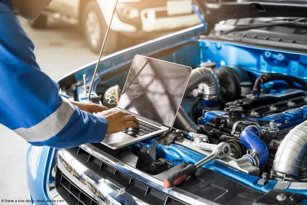 Ein Mechaniker arbeitet unter der offenen Motorhaube eines blauen Autos und hat Werkzeug im Motorraum.