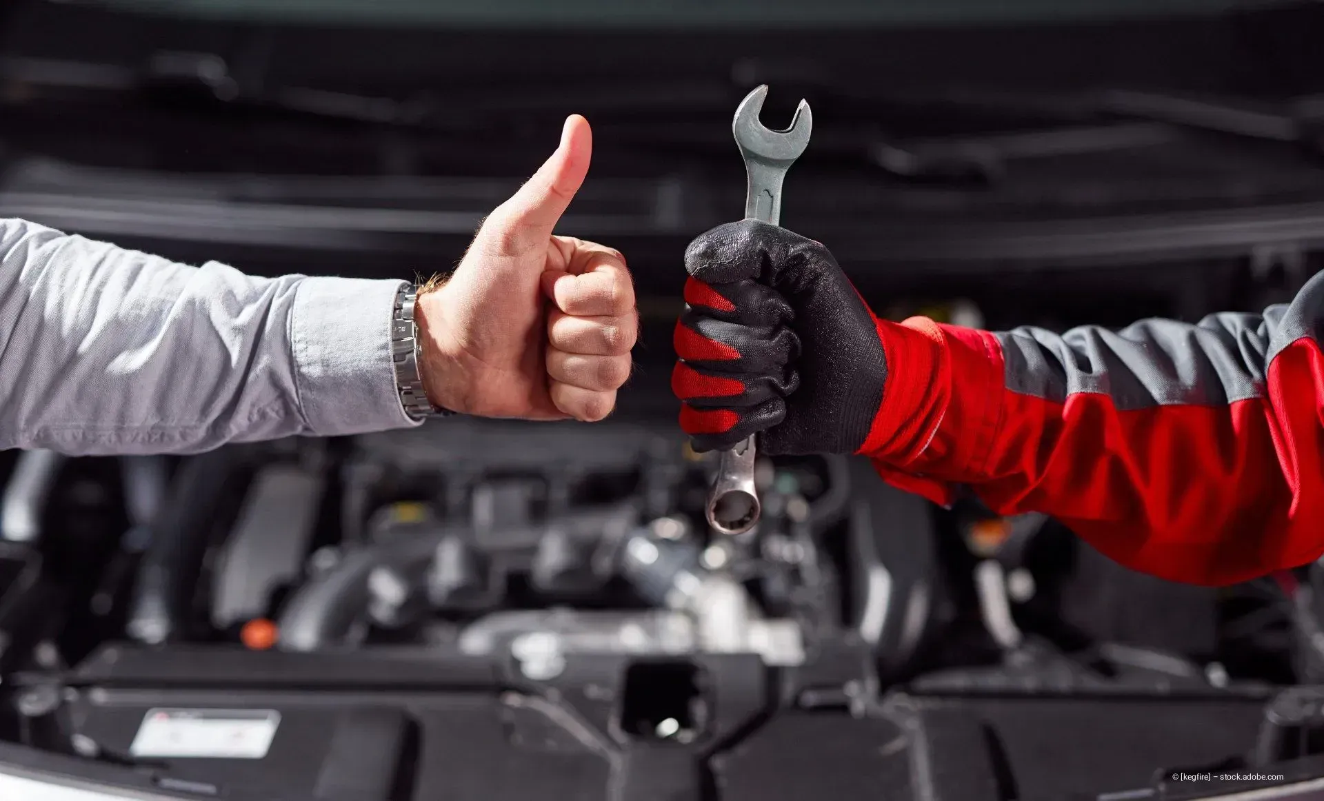 Zwei behandschuhte Hände: eine zeigt den Daumen nach oben, die andere hält einen Schraubenschlüssel vor einem Automotor.