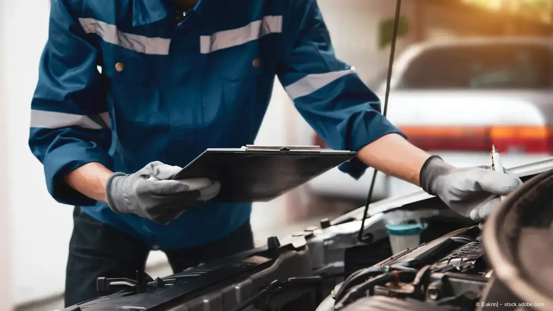 Ein Mechaniker in blauer Arbeitskleidung untersucht in einer Werkstatt mit einem Klemmbrett einen Automotor.