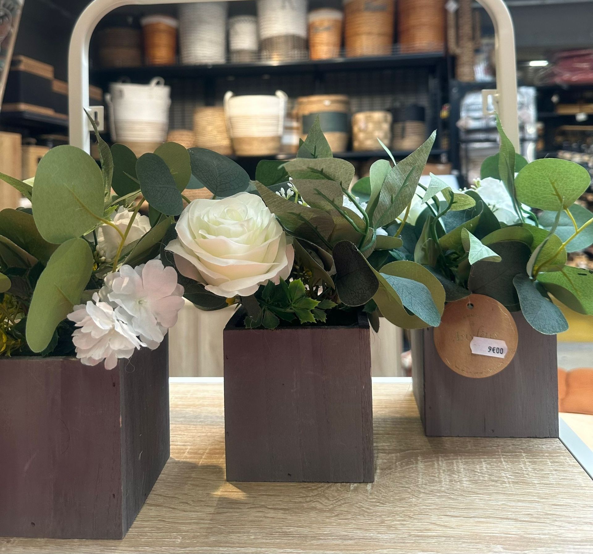 Des compositions de roses blanches et de verdure dans de petites boîtes en bois disposées sur une table, avec des étagères floues en arrière-plan.