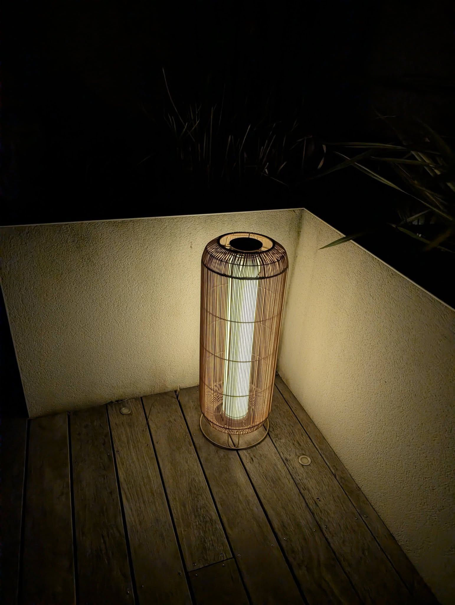 Une lanterne cylindrique éclairée, posée sur un plancher en bois dans un coin sombre.