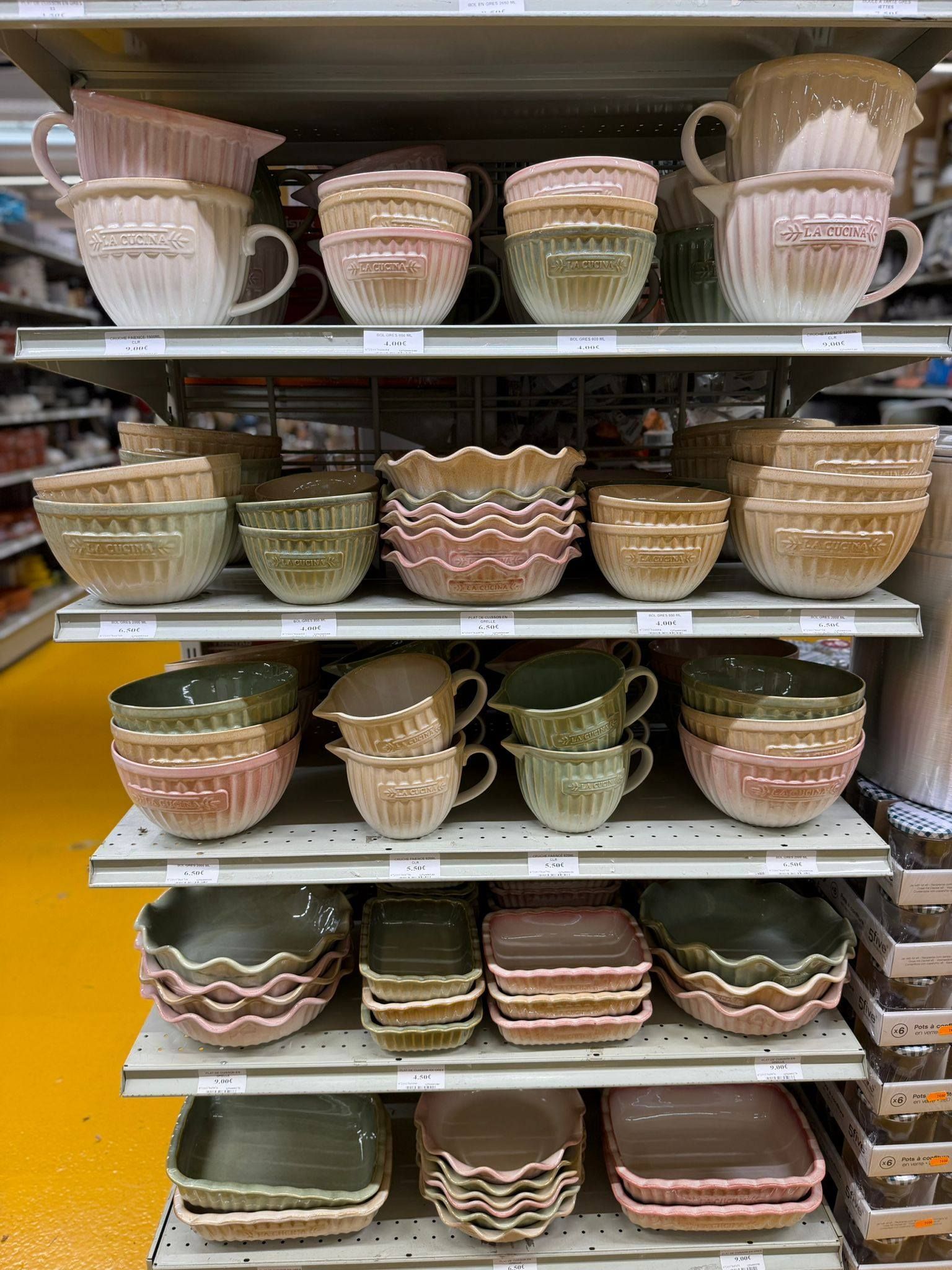 Des étagères remplies de bols et de plats décoratifs en céramique, dans des tons beiges, roses et verts, dans une allée de magasin.