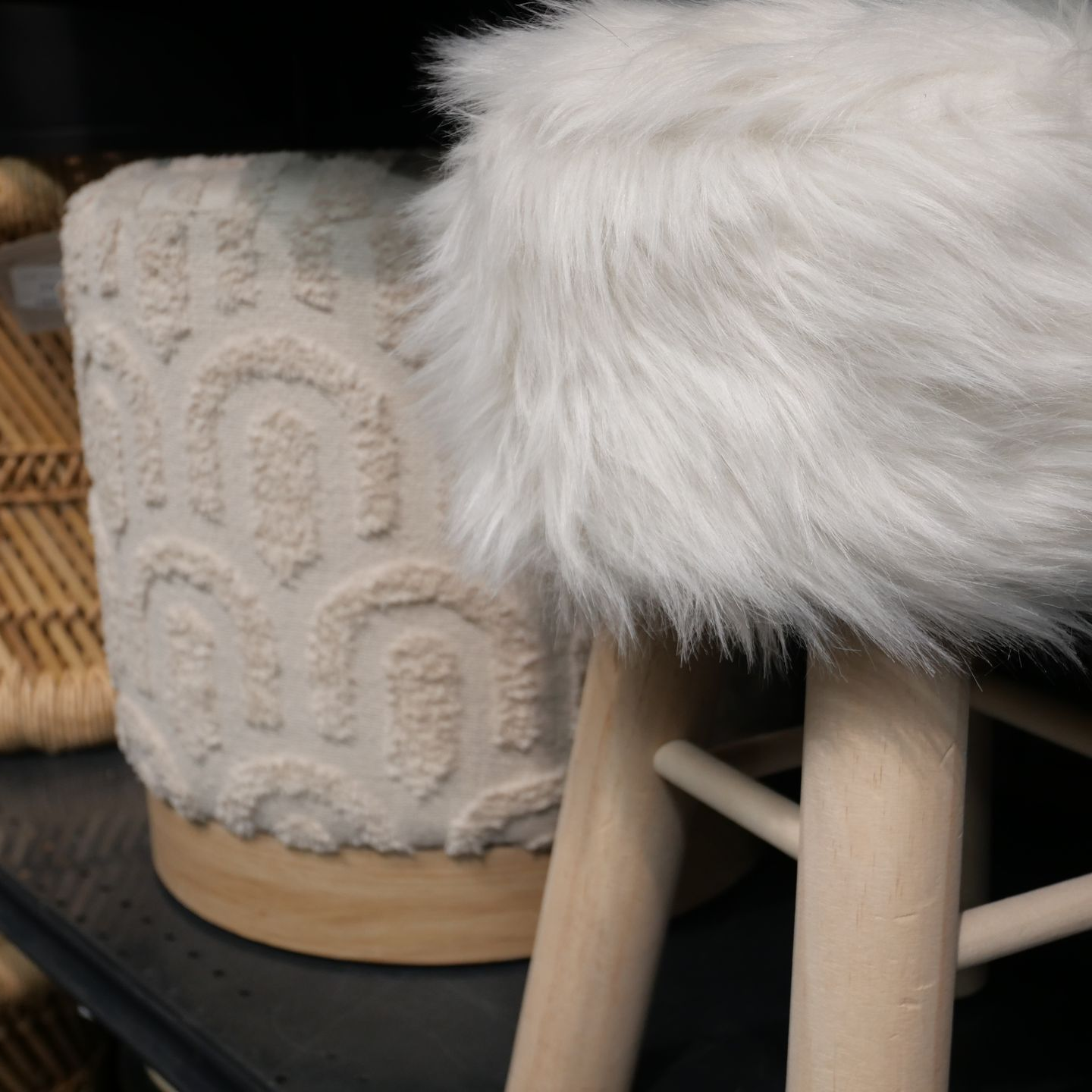 Housse de tabouret en fausse fourrure blanche sur un tabouret en bois clair, à côté d'un pouf beige tissé.