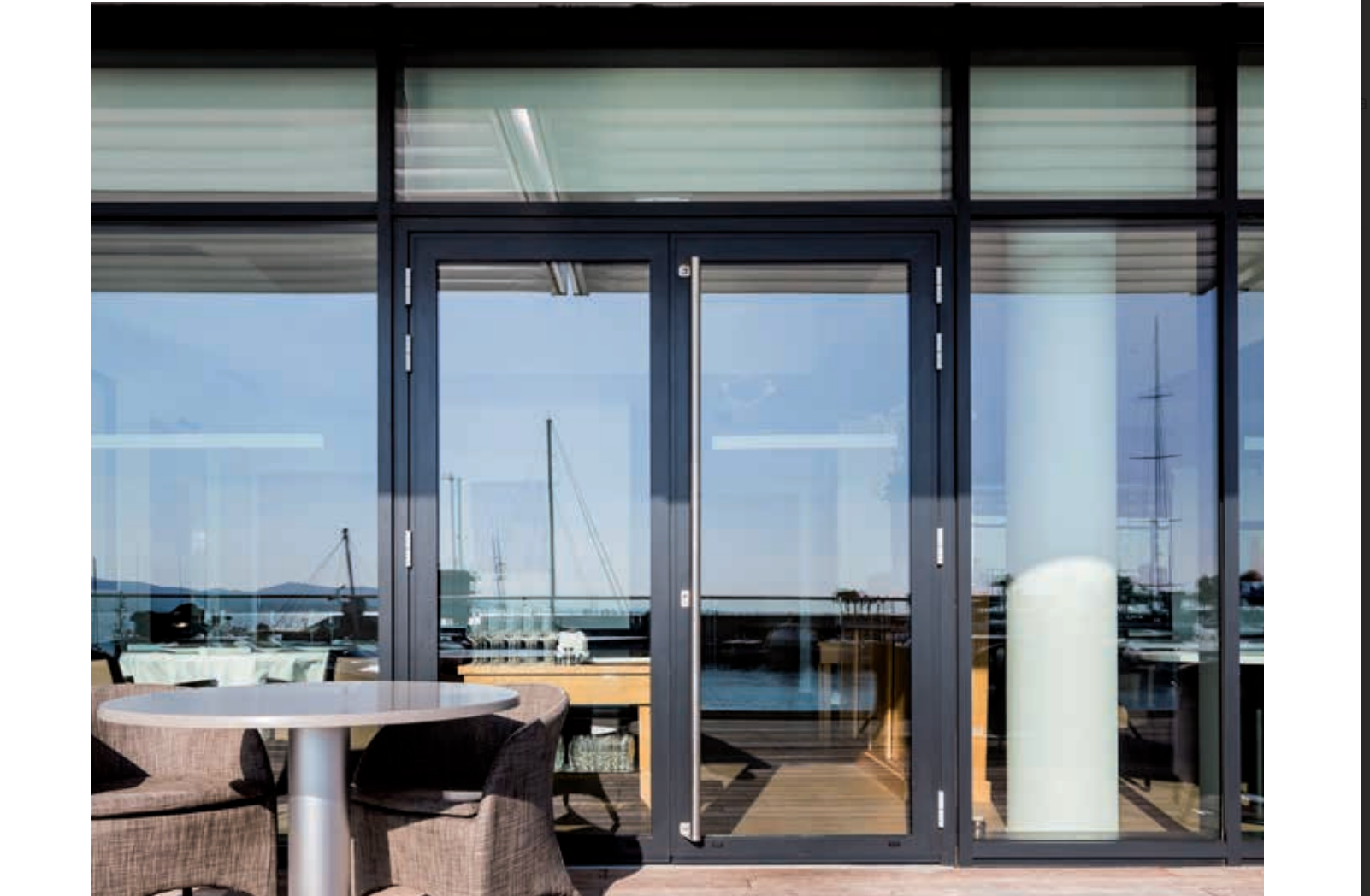 Les portes et fenêtres vitrées du restaurant donnent sur l'eau avec des bateaux, une table au premier plan.