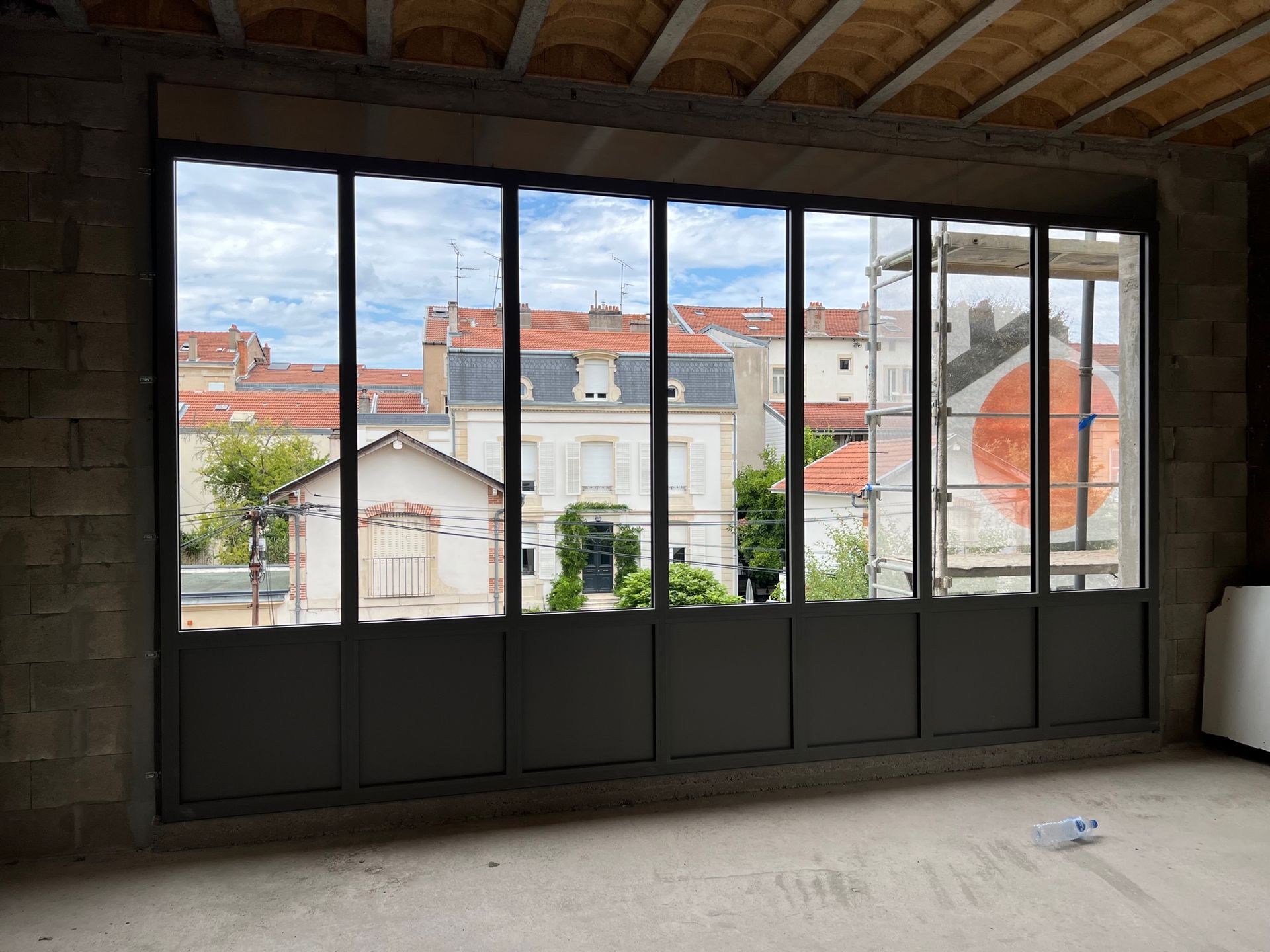 Grande fenêtre de style industriel à plusieurs vitres, donnant sur des bâtiments aux toits de tuiles rouges.