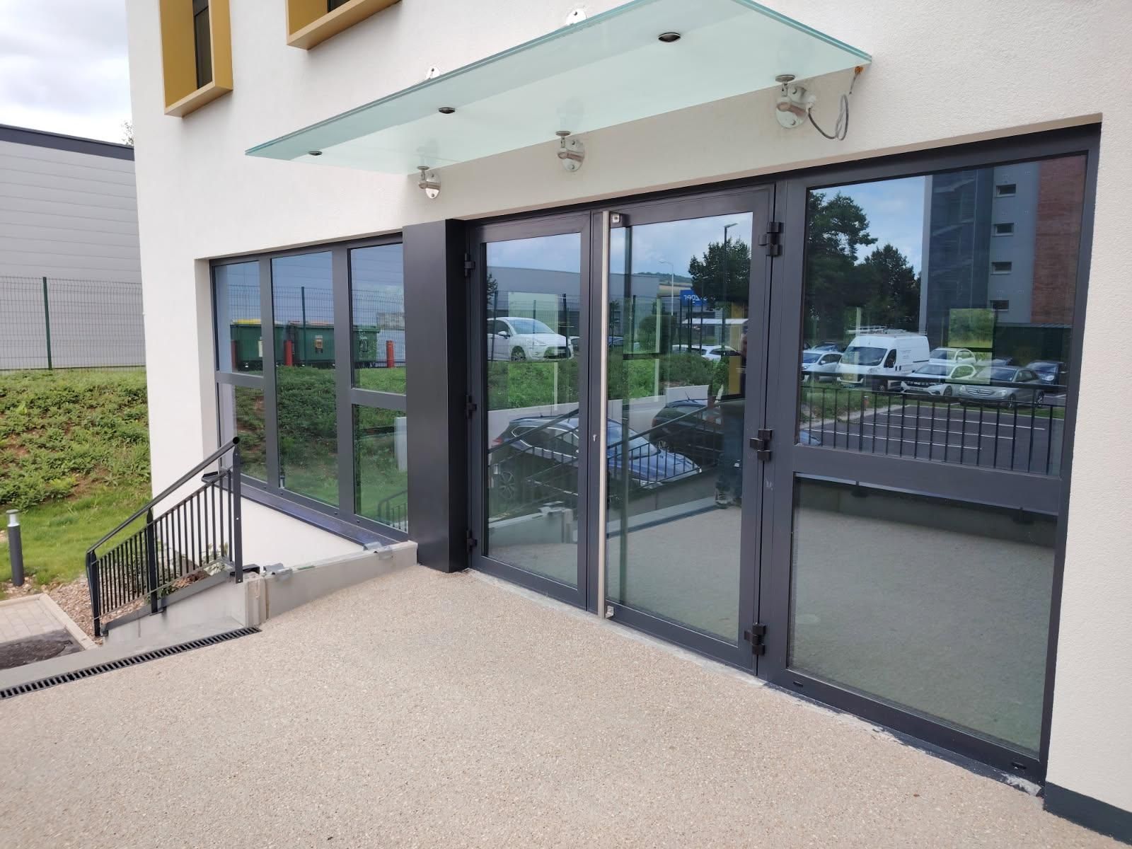Extérieur d'un bâtiment aux portes et fenêtres vitrées. Une rampe en béton mène à l'entrée. Un auvent en verre surplombe le bâtiment.