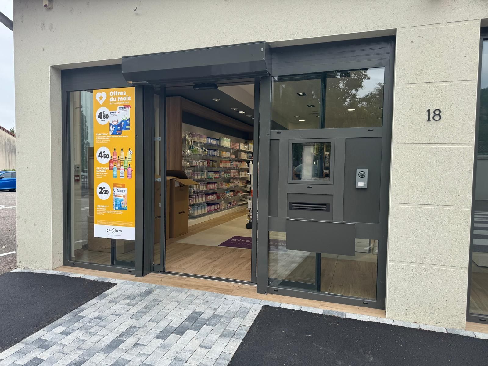 Entrée de la pharmacie avec porte ouverte, signalétique et distributeur automatique de billets. Façade du bâtiment portant le numéro 18.