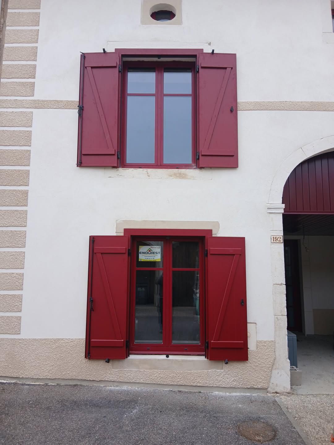 Volets rouges sur un bâtiment blanc avec fenêtres. Une porte cintrée est visible à droite.