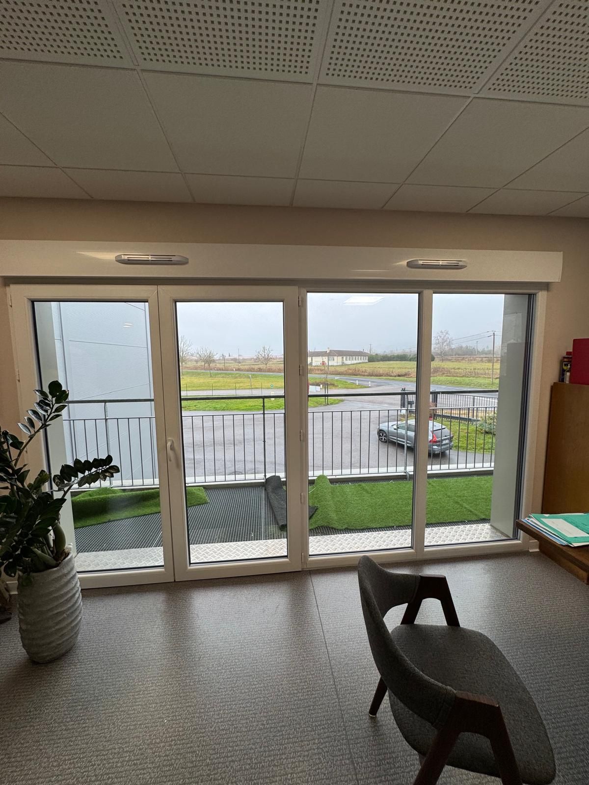 Des portes coulissantes en verre donnent sur un balcon, de la pelouse et une rue. Une chaise et une plante se trouvent dans la pièce.