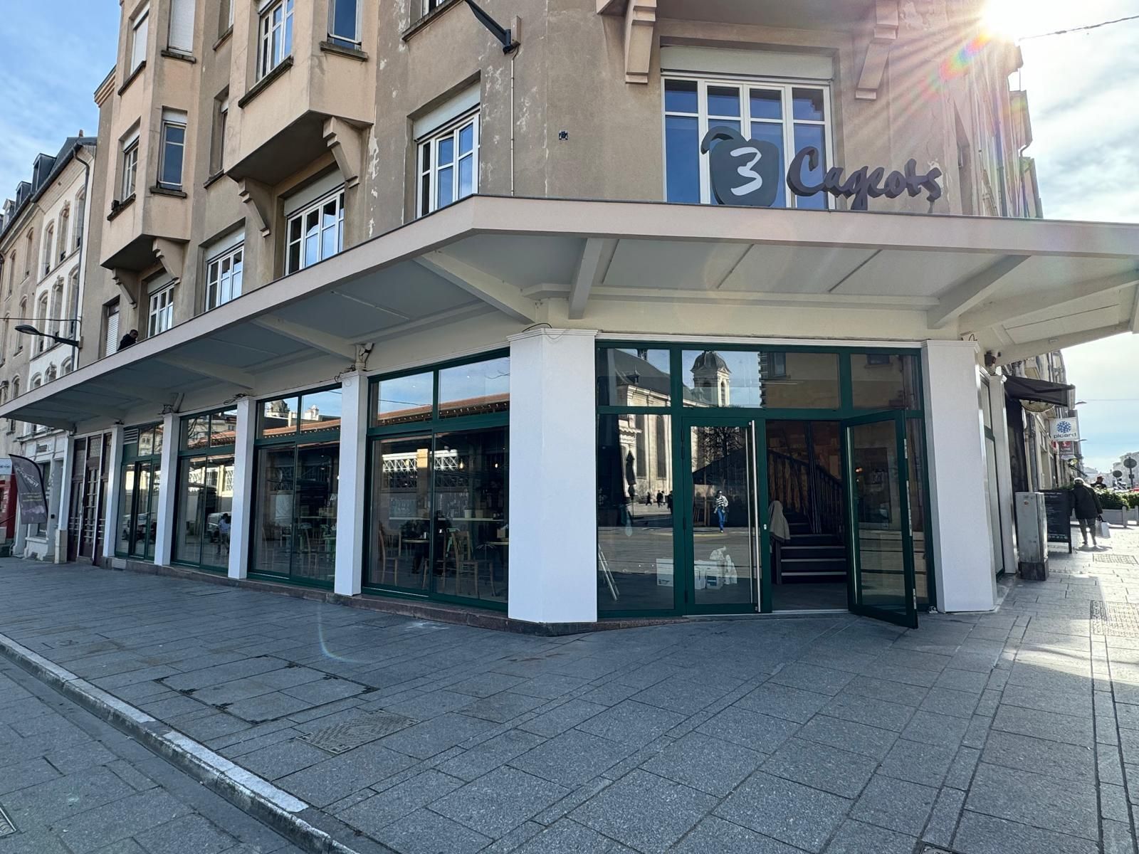 Le restaurant « 3 Cagots » est situé dans un immeuble d'angle avec des fenêtres en verre, un auvent blanc et une vue sur la rue.