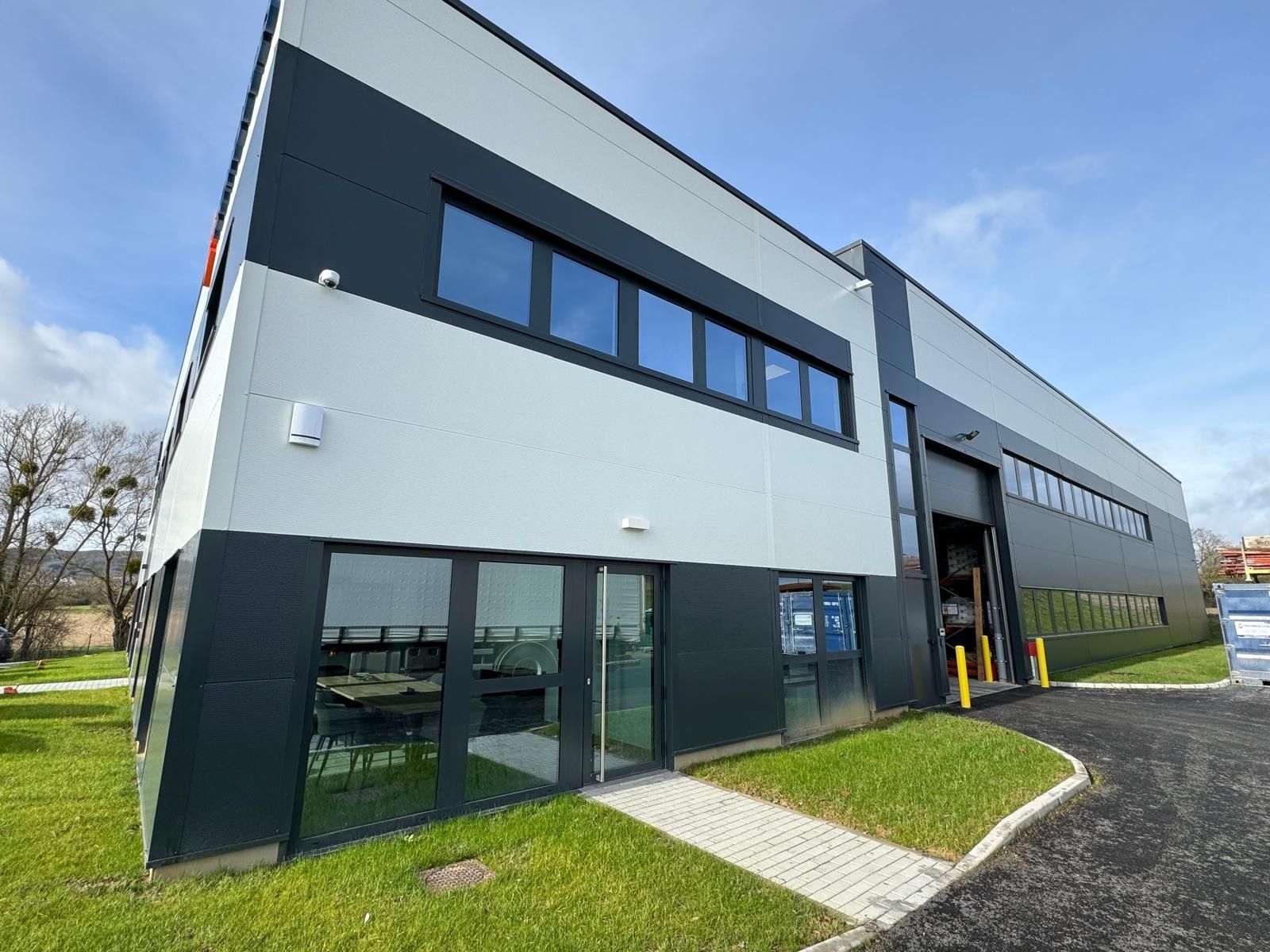 Bâtiment industriel moderne de deux étages avec panneaux noirs et blancs, fenêtres en verre et entrée pavée.