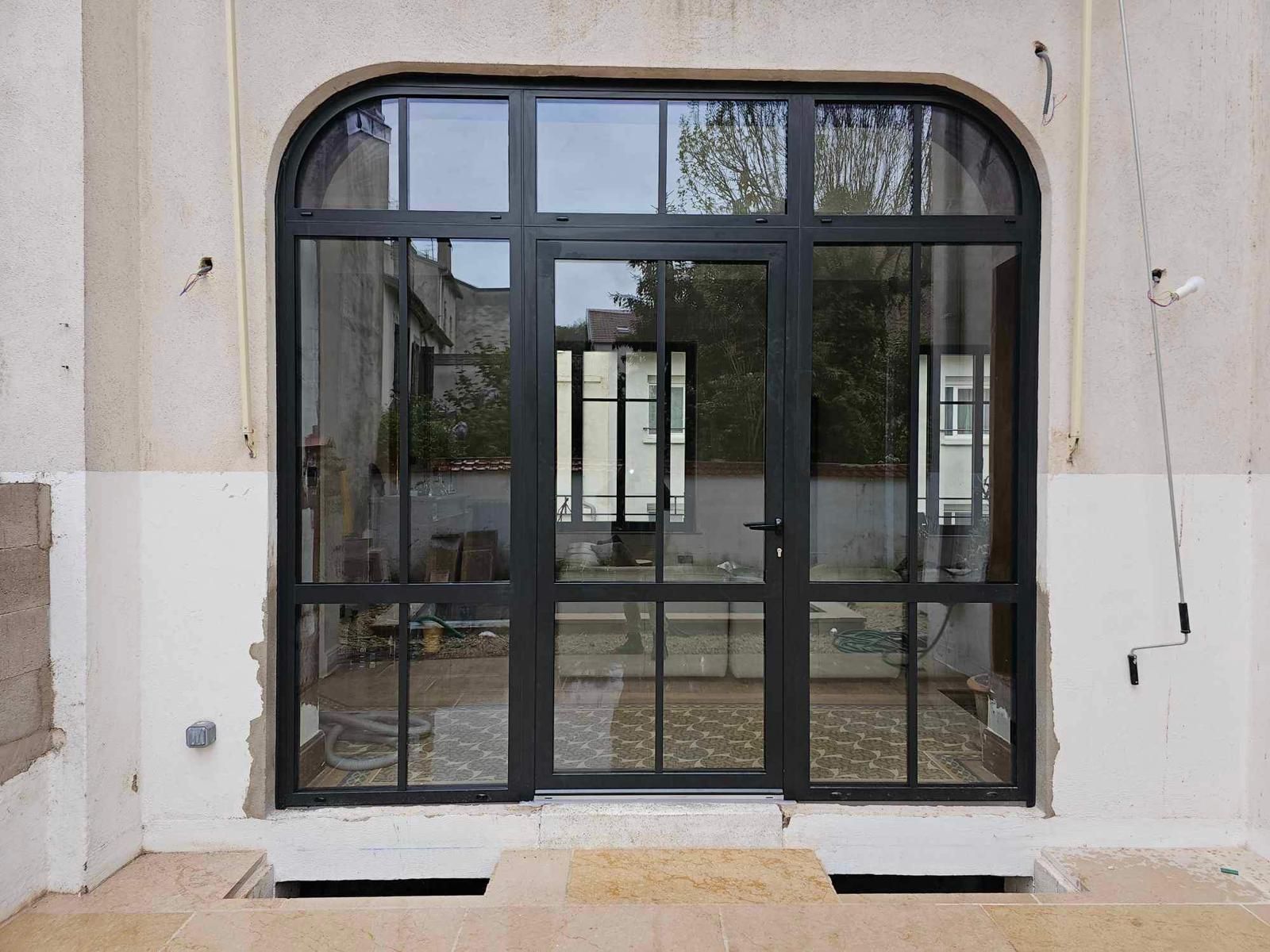 Porte gris foncé avec fenêtre rectangulaire au-dessus, longue poignée argentée, encadrement en pierre beige. Numéro 15 au-dessus.