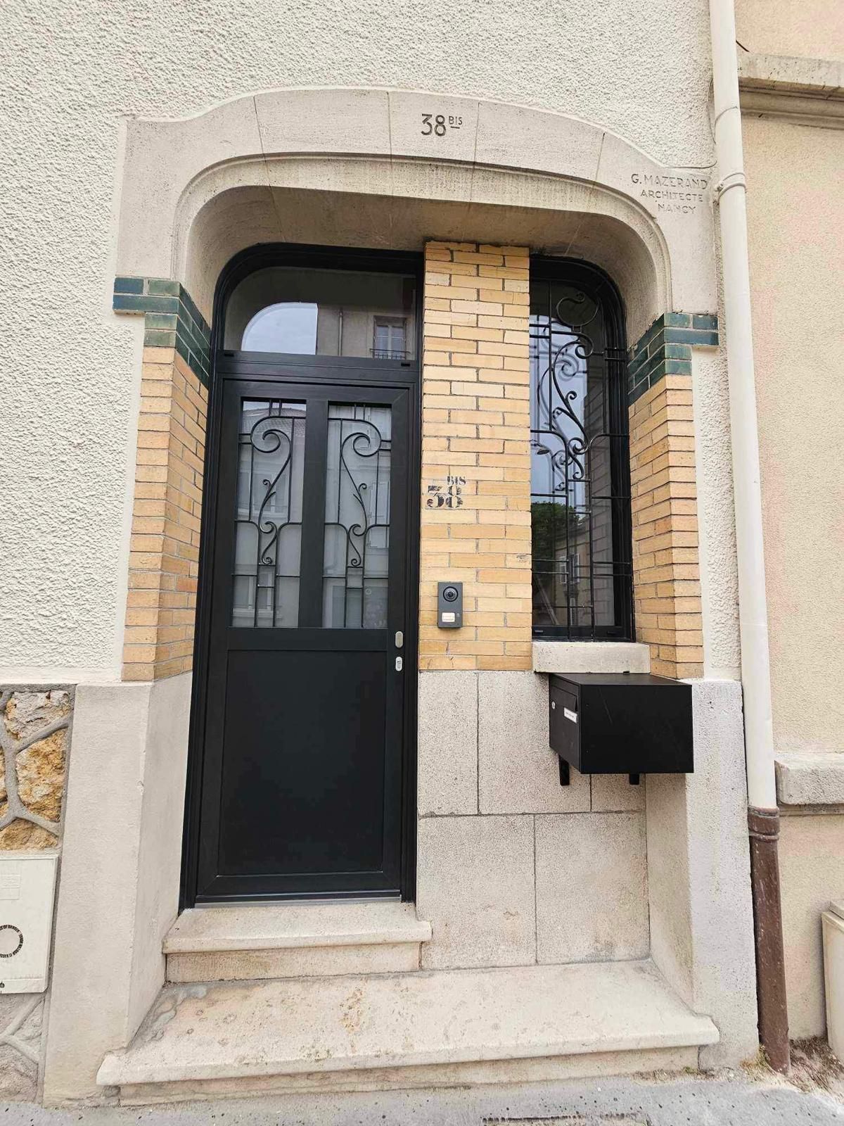 Porte noire ornée de ferronneries décoratives, fenêtre et boîte aux lettres sur un bâtiment en briques et en pierres.