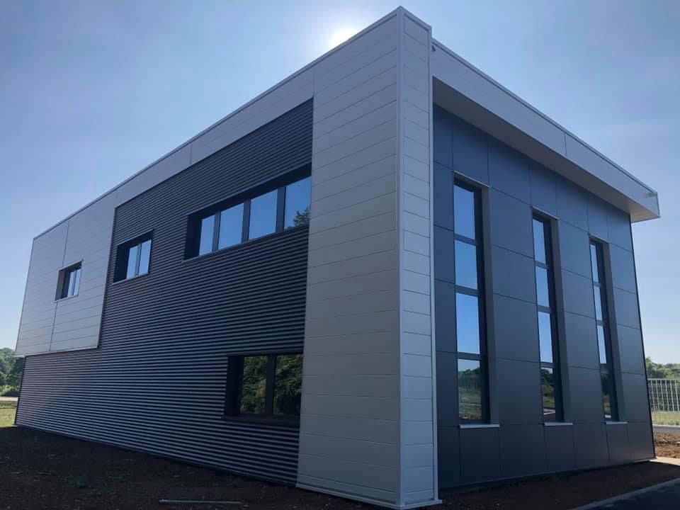 Portes vitrées bleues avec poignée argentée. Une personne se tient entre les portes, son reflet est visible. Voitures garées et un bâtiment en arrière-plan.