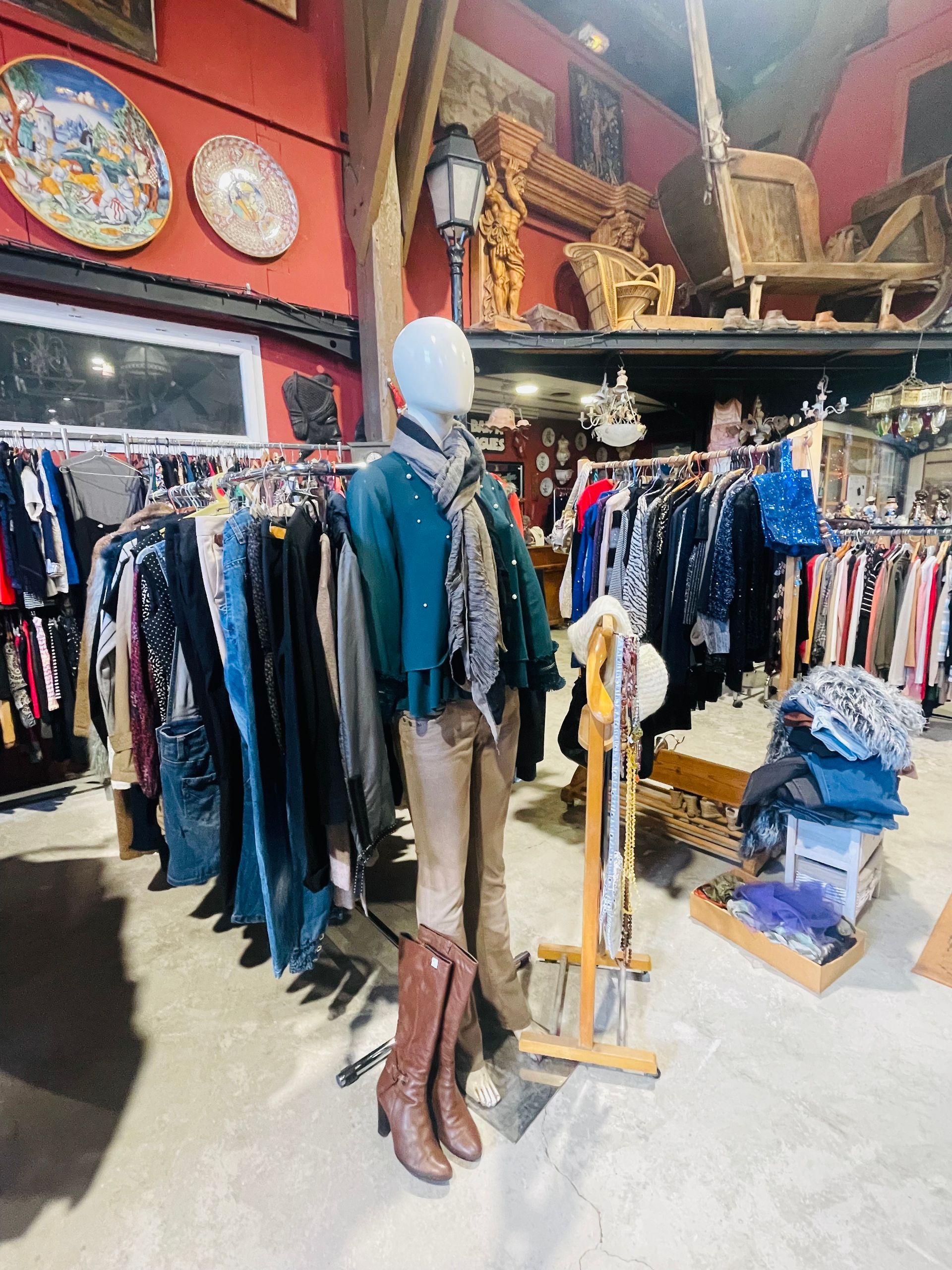 Un mannequin portant une veste verte, une écharpe et des bottes marron se tient dans l'allée d'un magasin de vêtements rempli de portants.