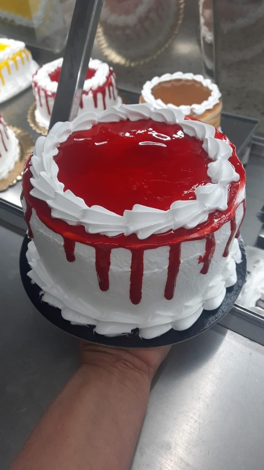 Pastel con glaseado blanco y rojo, sostenido por una mano. Otros pasteles al fondo.