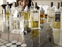 Mulher rodeada por frascos de perfume, observando atentamente as fragrâncias sobre uma mesa.