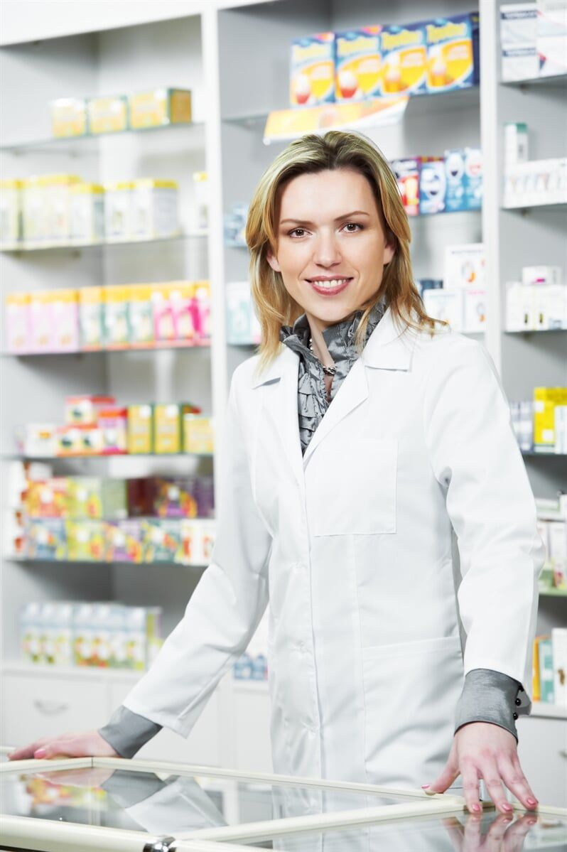 Farmacêutico de jaleco branco sorri atrás do balcão de uma farmácia. Prateleiras com produtos estão visíveis.