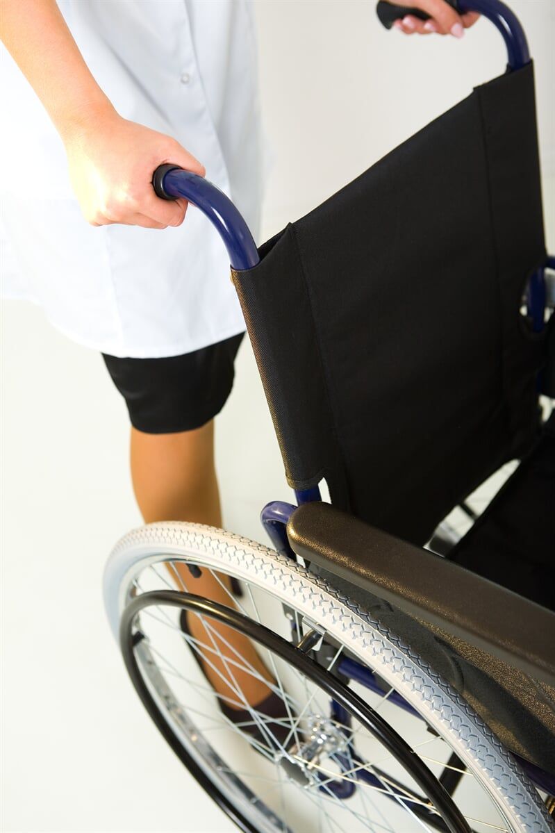 Pessoa de uniforme branco empurrando uma cadeira de rodas com assento preto e alças azuis.