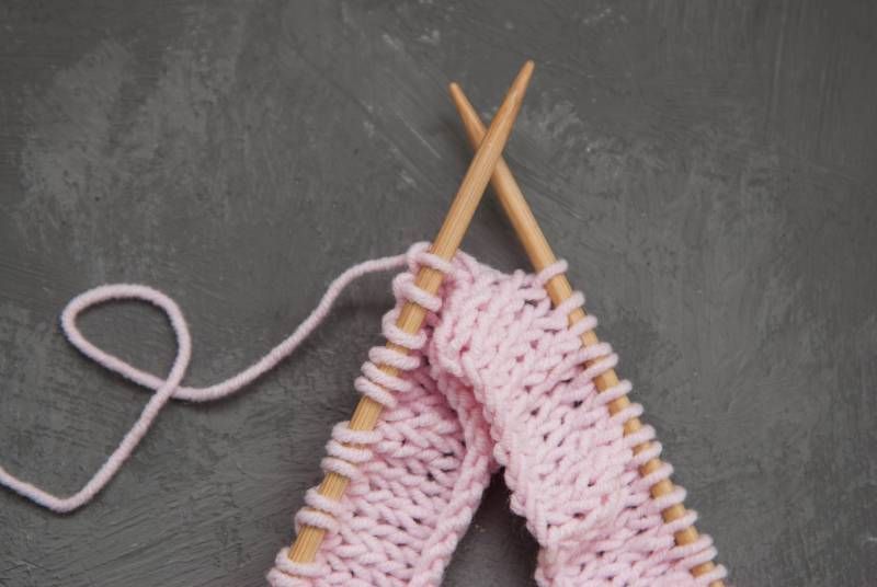 zwei überkreuzte Stricknadeln mit rosa Wolle und angefangenem Projekt auf grauem Hintergrund