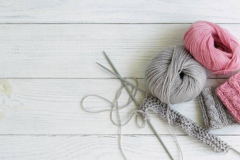 auf weißem Holz liegen auf der rechten Seite graue und rosa Wolle mit Stricknadeln und einem angefangenen Projekt
