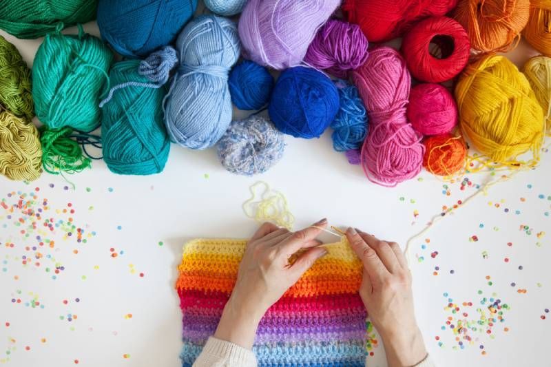 zwei Hände stricken ein buntes Stück und vor dem Händen ist ein Haufen in einer Reihe angeordnet mit Wolle in allen Regenbogen Farben