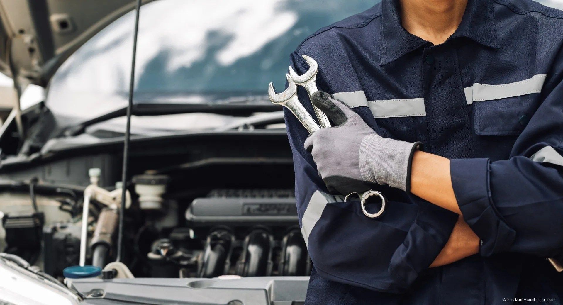 Ein Mechaniker hält Schraubenschlüssel neben einer offenen Motorhaube in einer Werkstatt.