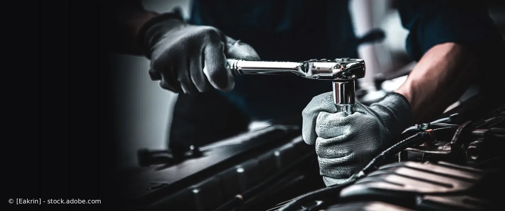 Ein Mechaniker zieht in einer dunklen Werkstatt mit schwarzen Handschuhen ein Autoteil mit einer Ratsche fest.