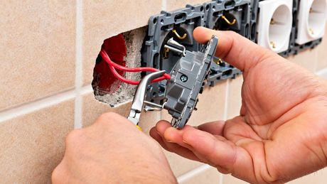 Unas manos conectando un enchufe eléctrico en una pared de azulejos, utilizando cables rojos y un destornillador.