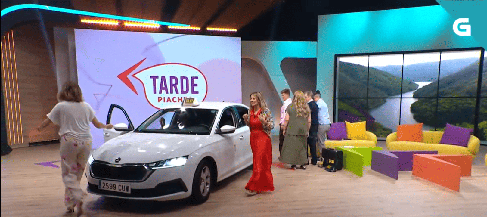 Un estudio de televisión con un coche plateado, gente de pie cerca, sillas coloridas y un fondo con una ventana panorámica.