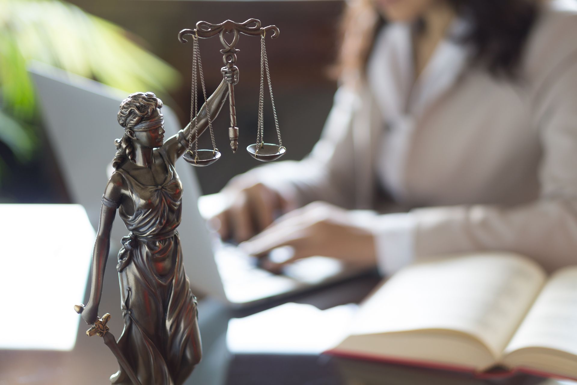 Statue de la justice au premier plan, avec un avocat flou travaillant à son bureau et un livre de droit ouvert en arrière-plan.