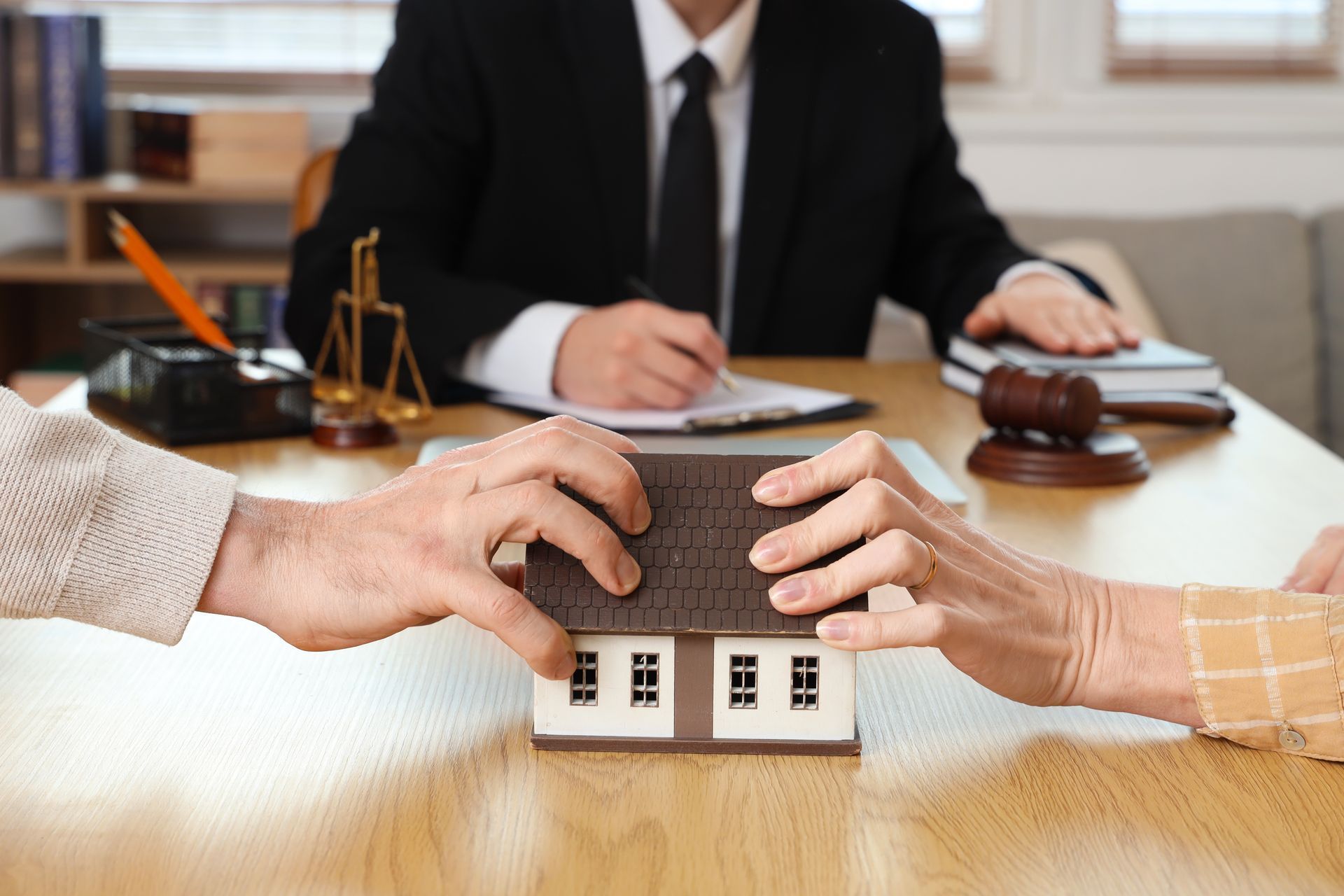 Des mains échangent une maquette de maison à un bureau lors d'une transaction immobilière, un avocat étant visible en arrière-plan.
