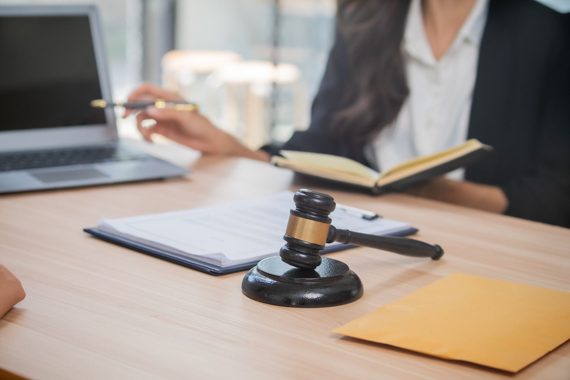 Bureau d'avocat avec marteau, ordinateur portable et carnet ; une personne assise à l'arrière-plan tient un stylo.