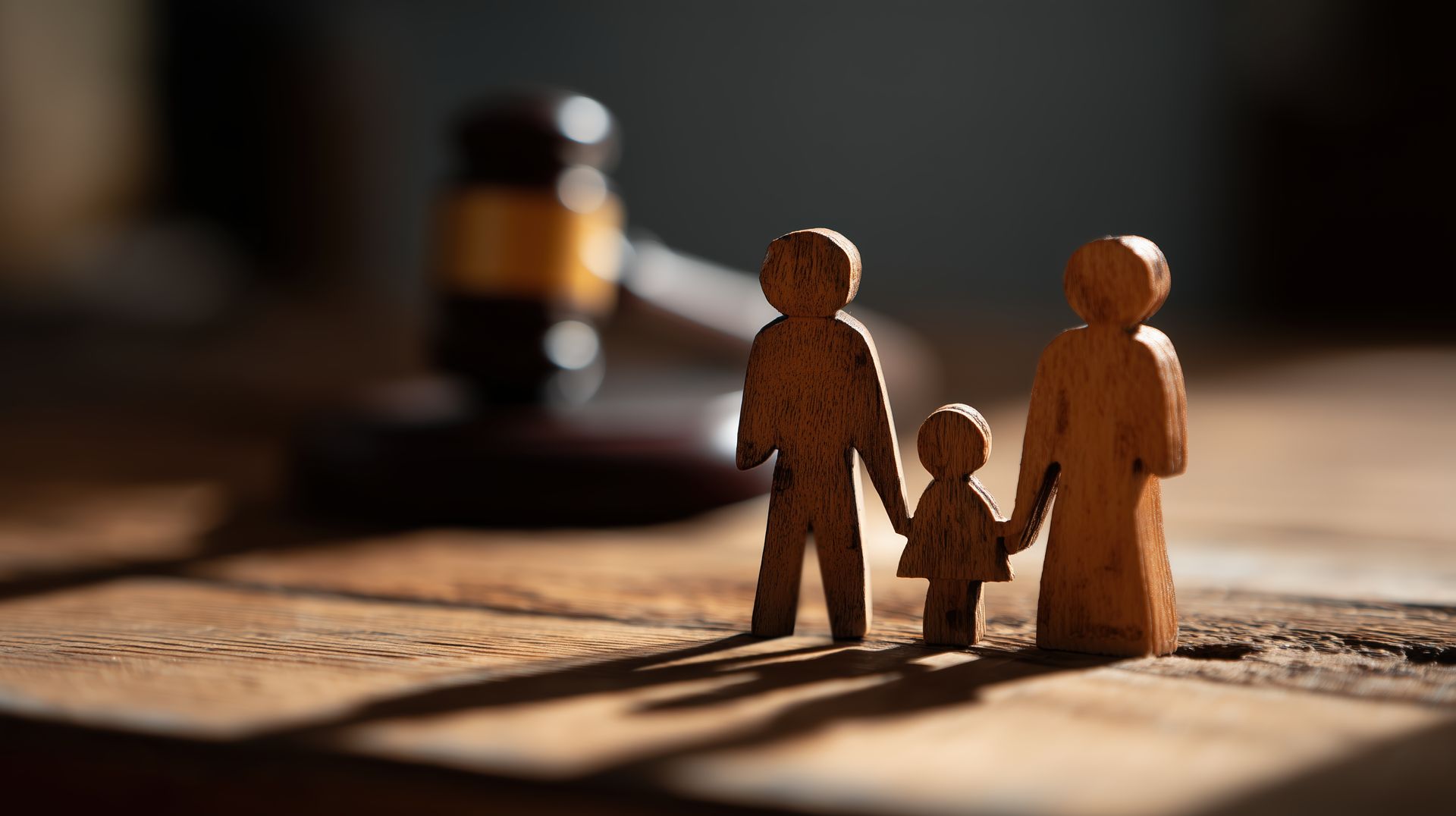 De petites figurines en bois représentant une famille se tenant la main sont posées sur une table ensoleillée, avec un bocal en arrière-plan flou.