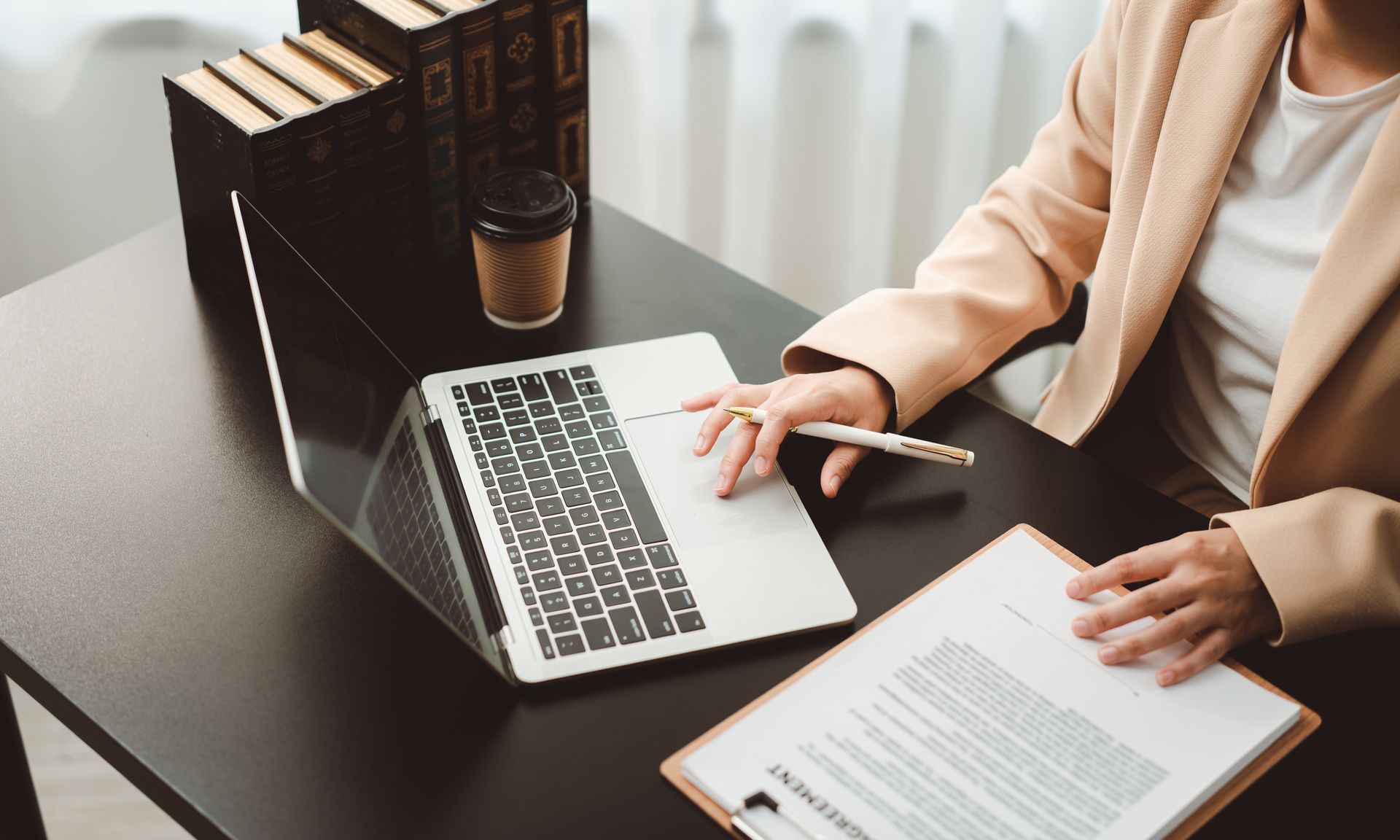 Une personne travaille sur un ordinateur portable, avec des cahiers et une tasse de café sur un bureau.