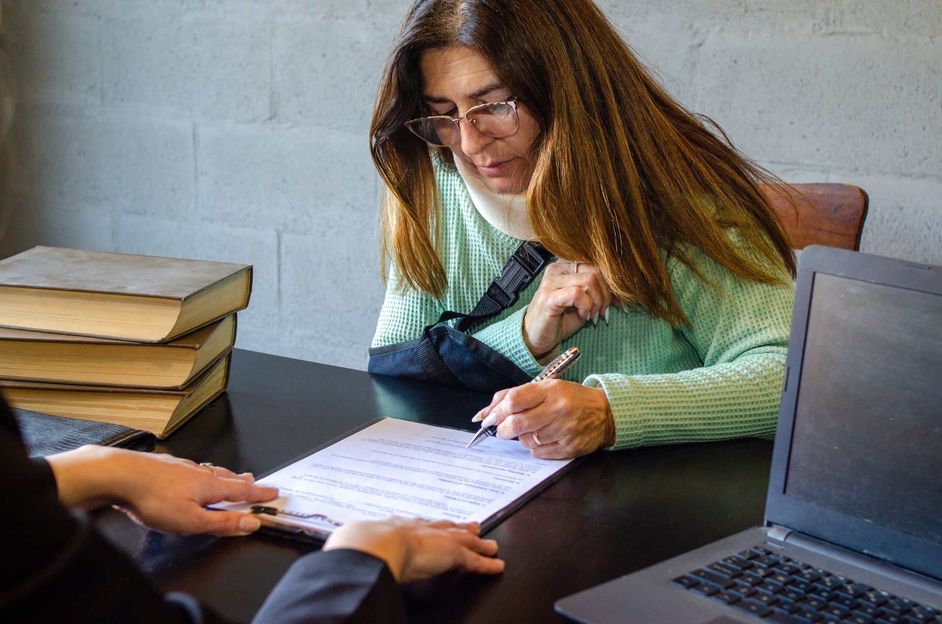 Une femme examine des documents à une table avec un ordinateur portable et des livres empilés à proximité.