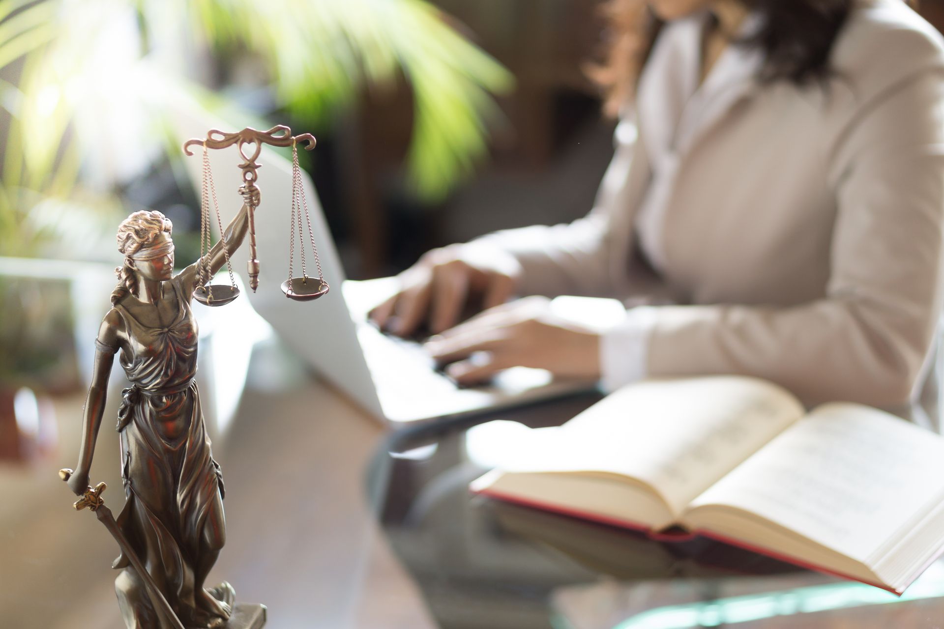 Un livre de droit et une balance au premier plan, une personne tapant sur un ordinateur portable dans un bureau lumineux