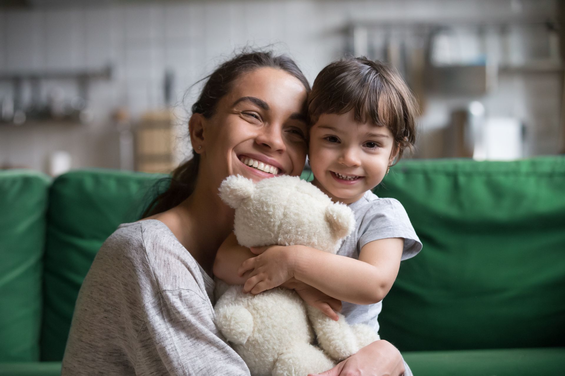 Un adulte et un enfant souriants, assis sur un canapé vert, enlaçant un ours en peluche blanc dans un salon confortable.