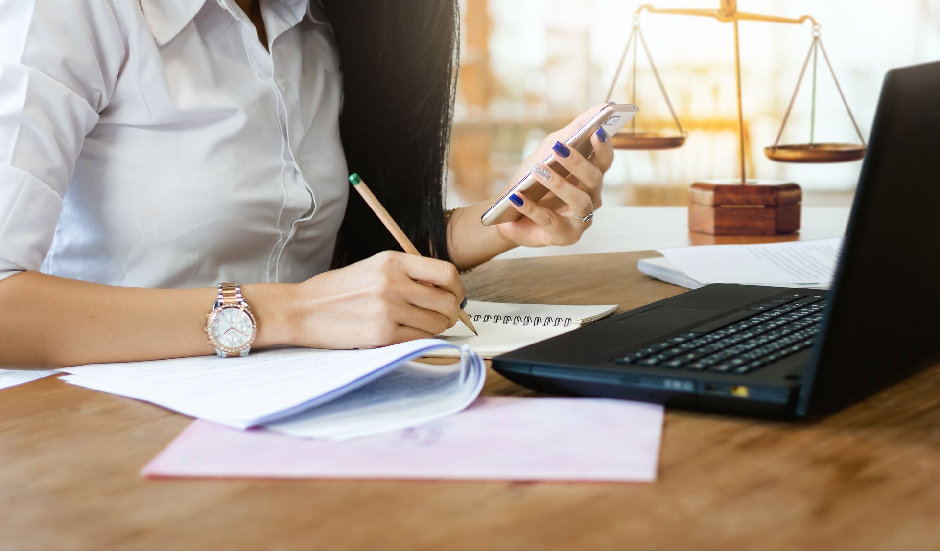 Une personne consulte des documents et un smartphone à côté d'un ordinateur portable sur un bureau, avec une balance judiciaire en arrière-plan.