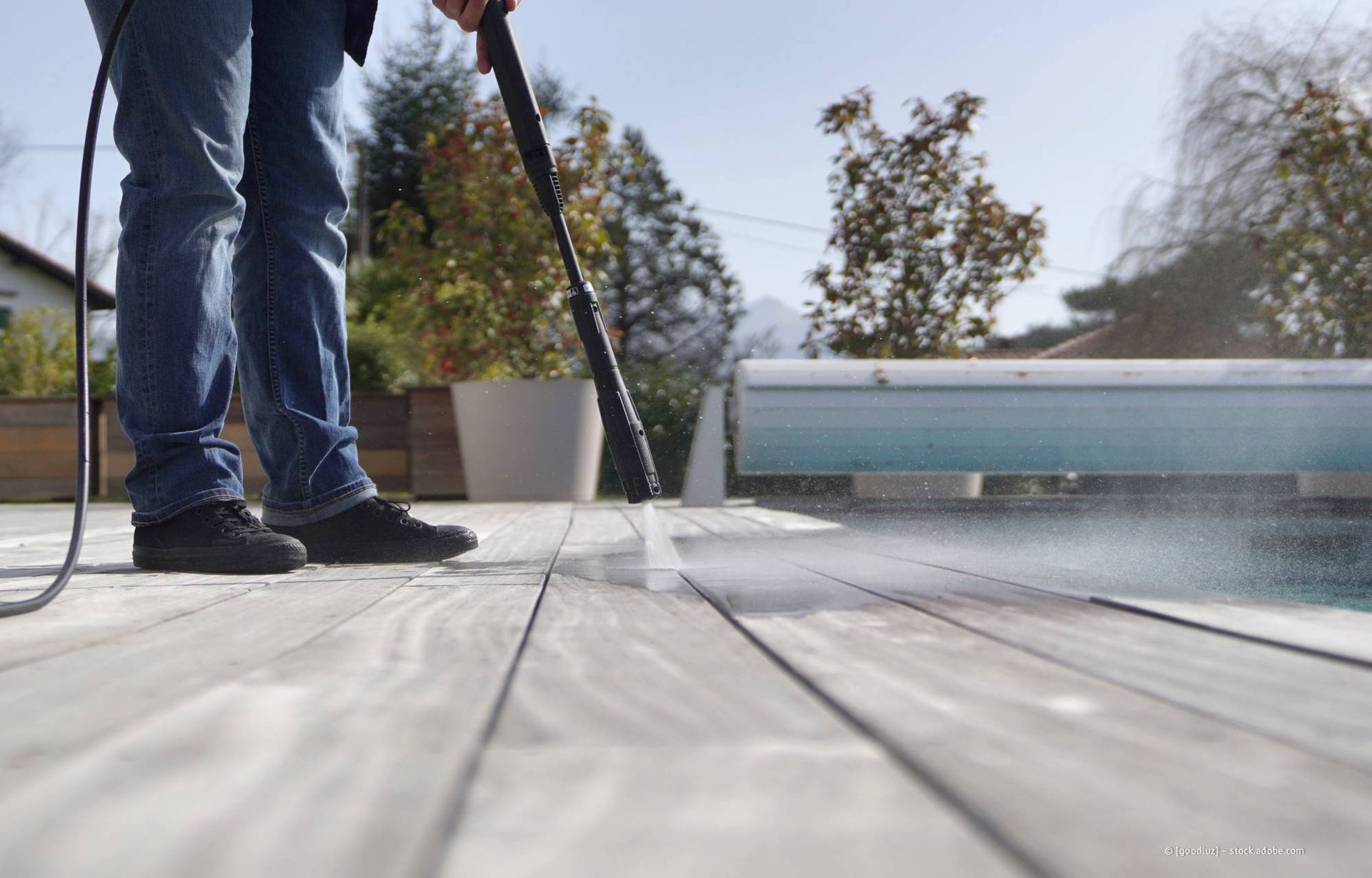 Eine Person reinigt an einem sonnigen Tag mit einem Hochdruckreiniger eine Holzterrasse in der Nähe eines Pools.