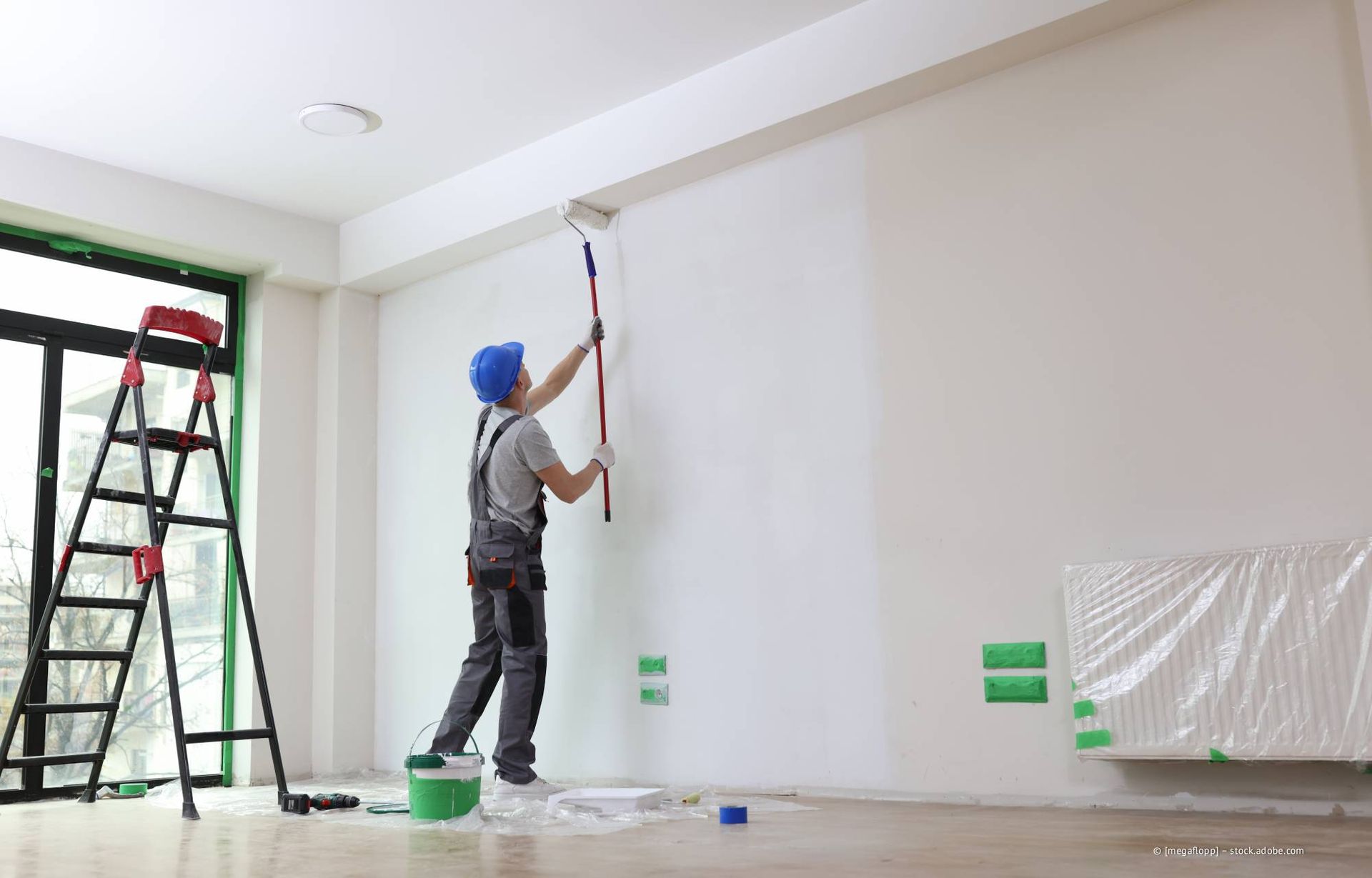 Ein Maler mit blauem Schutzhelm streicht mit einer Farbrolle eine weiße Wand. Leiter und Farbeimer stehen daneben.