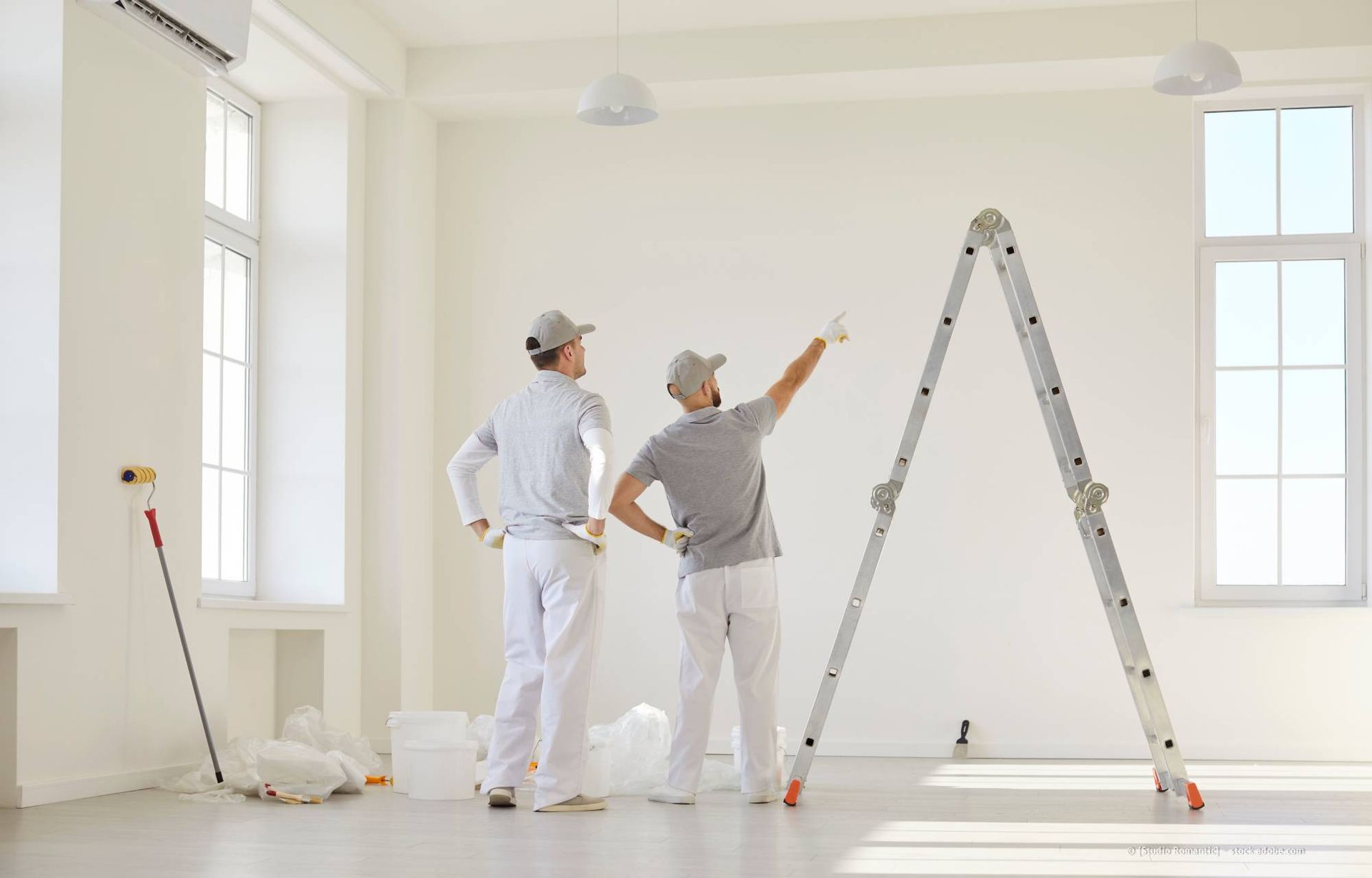 Zwei Maler in einem Raum mit einer Leiter, einer zeigt auf eine Wand. Weiße Farbe, lichtdurchfluteter Raum.
