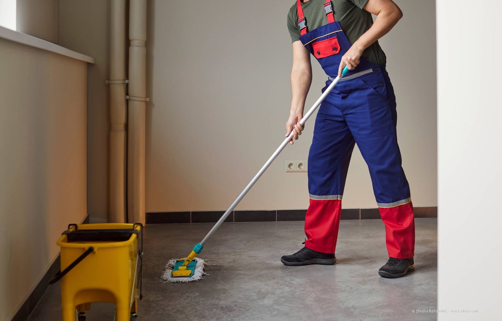 Eine Person in blauem Overall wischt einen Betonboden neben einem gelben Eimer.