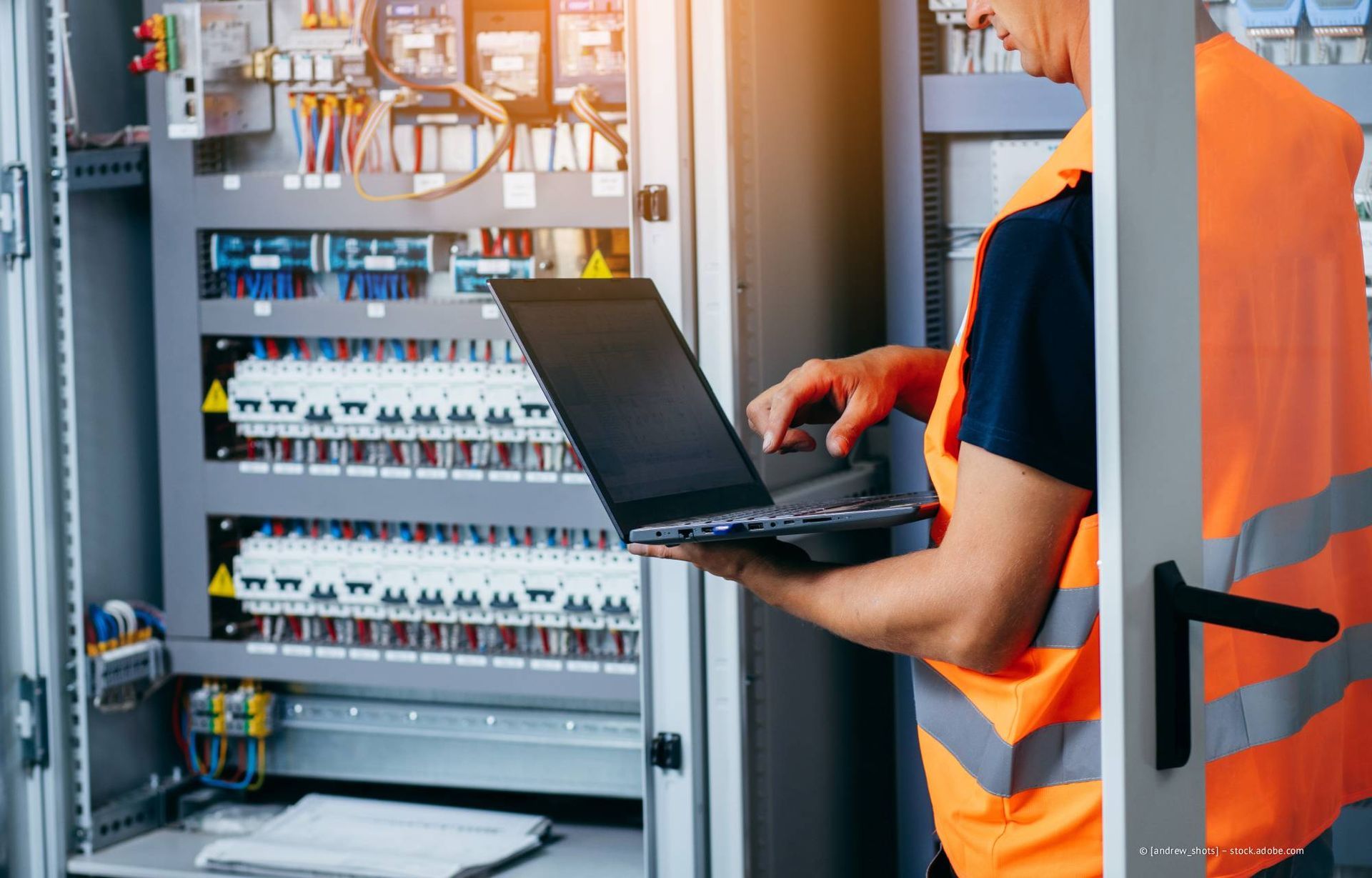 Elektriker benutzt Laptop vor einem Schaltschrank; er trägt eine Sicherheitsweste.
