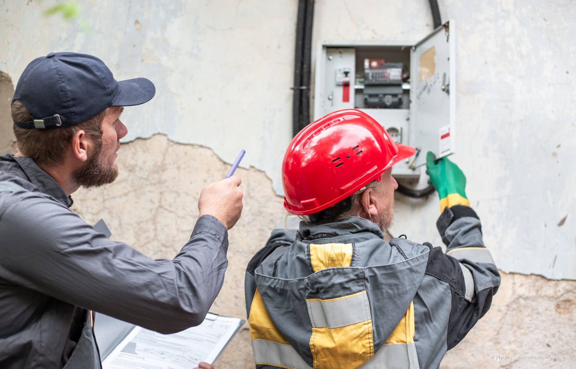 Zwei Arbeiter inspizieren einen Elektrokasten an einer verwitterten Wand; einer zeigt darauf, der andere trägt einen Schutzhelm und Handschuhe.