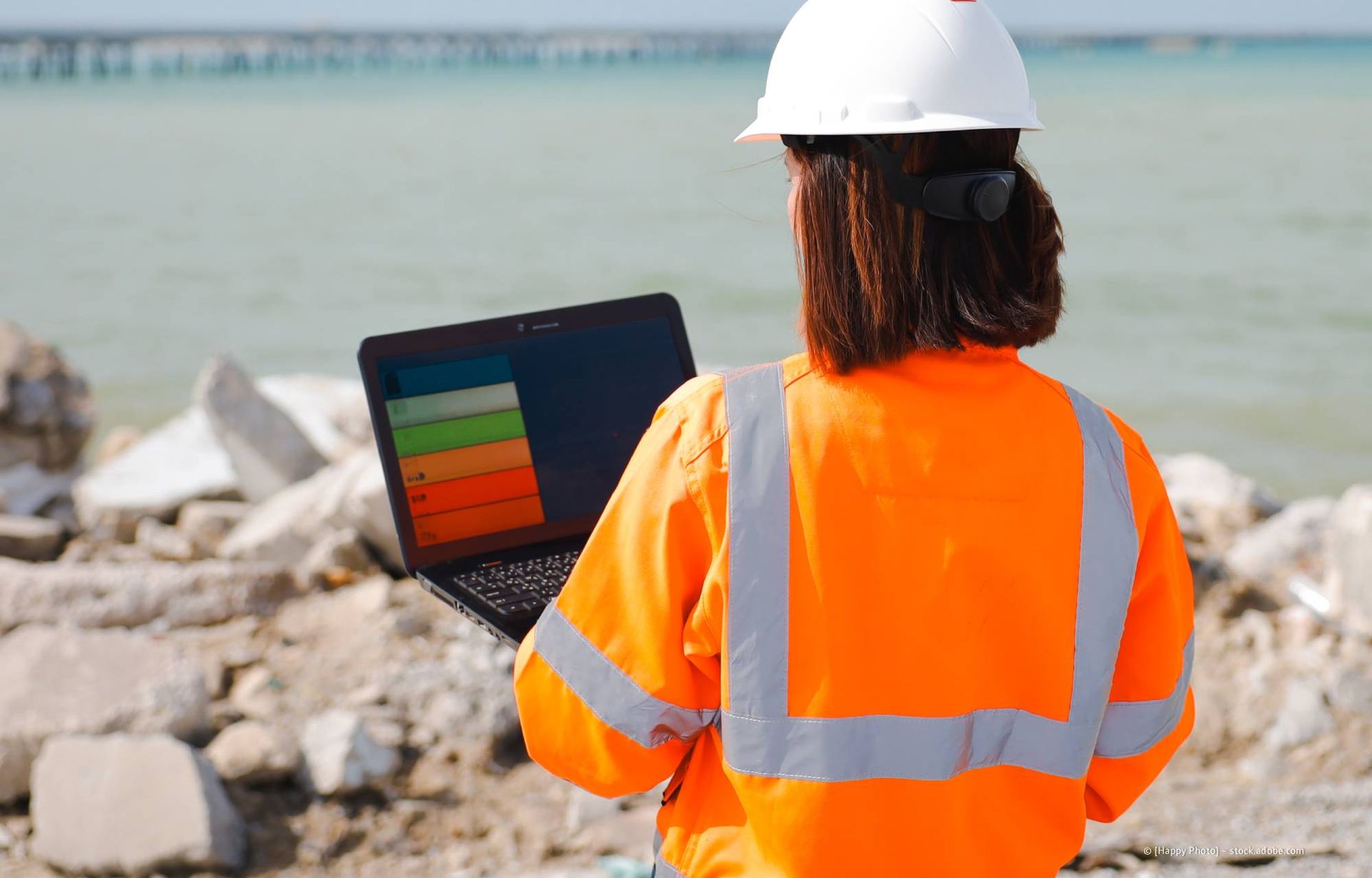 Eine Person in orangefarbener Arbeitskleidung und Schutzhelm schaut im Freien in der Nähe von Wasser und Schutt auf einen Laptop-Bildschirm.