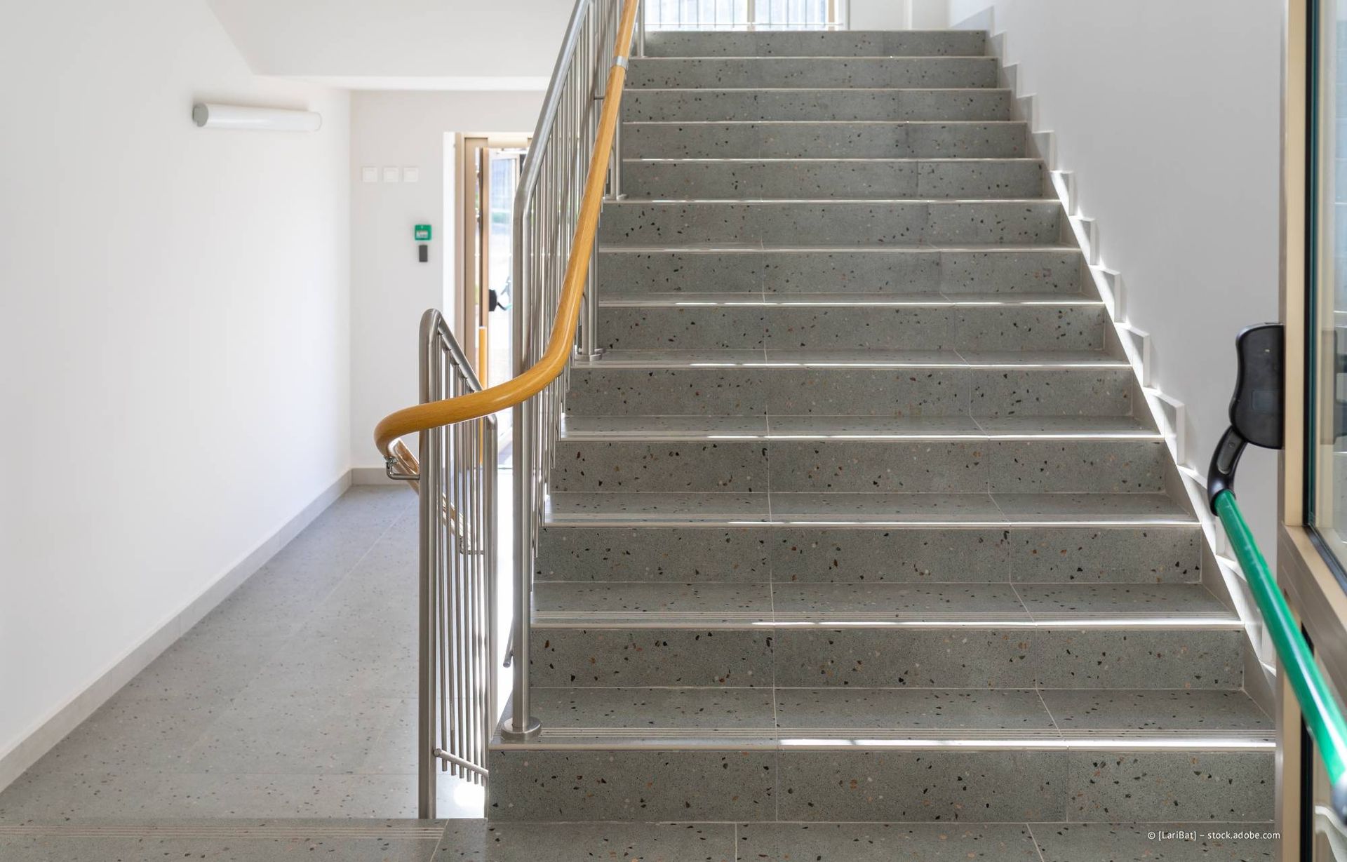 Treppe mit grau gesprenkelten Stufen, Metall- und gelben Handläufen, weißen Wänden und einem Korridor.