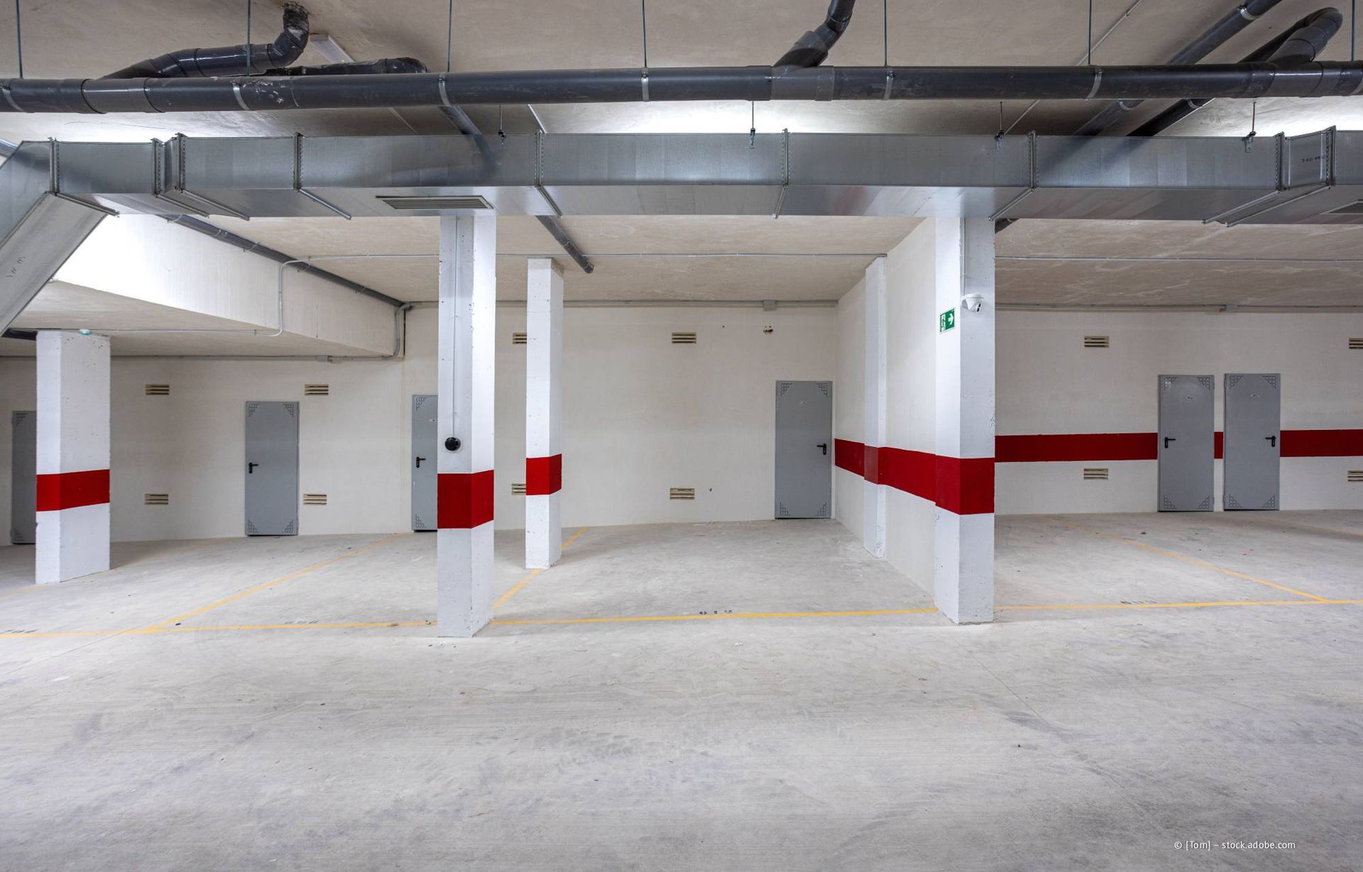 Leeres Parkhaus mit weißen Säulen und roten Streifen. Graue Türen und Lüftungskanäle.