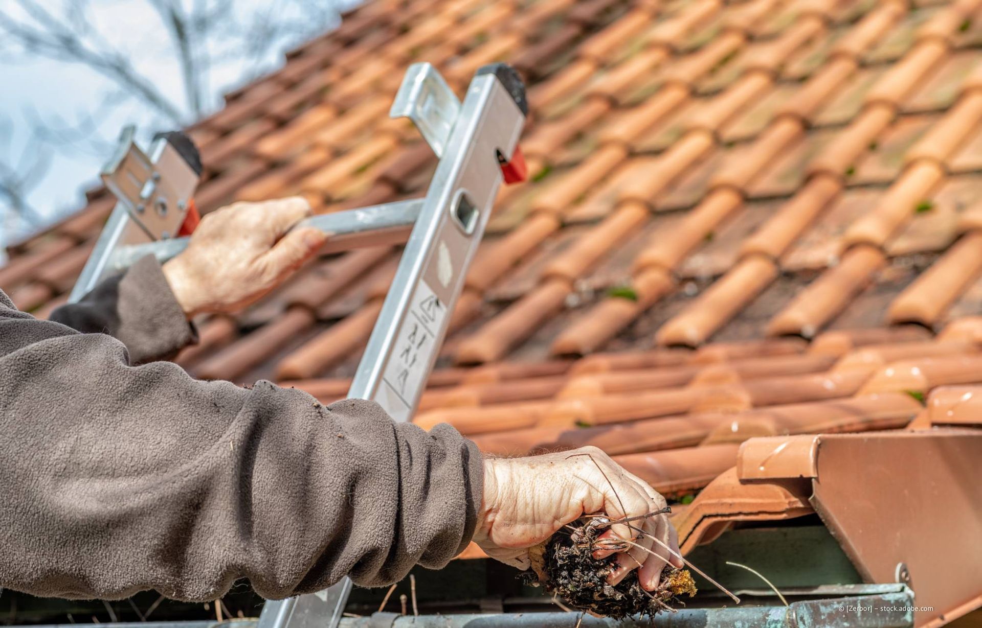 Eine Person auf einer Leiter reinigt eine mit Ablagerungen von einem roten Ziegeldach gefüllte Dachrinne.